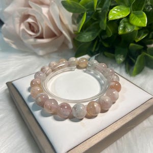 Puede incluir: Una pulsera de cuentas con piedras preciosas redondas de color rosa claro y melocotón. La pulsera se exhibe sobre una superficie blanca dentro de una caja dorada, con un fondo borroso de una rosa rosa y follaje verde.
