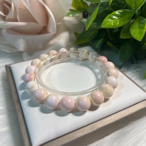 May include: A bracelet made of round, pale pink and white beads. The bracelet is presented on a white surface, with a blurred backdrop of a pink rose and green foliage. The beads are approximately 1.3 cm in diameter.