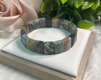 May include: A multi-colored gemstone bracelet with rectangular beads. The bracelet features shades of green, gray, and purple. The bracelet is displayed on a white surface with a neutral-toned border, with a rose and greenery in the background.