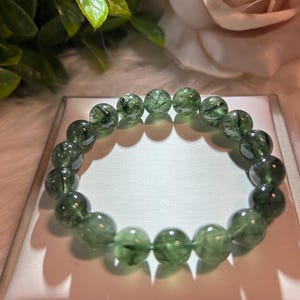 May include: A green beaded bracelet featuring translucent, round beads with a speckled, web-like pattern. The bracelet is displayed on a white surface, with a blurred background of green leaves and a pink rose.