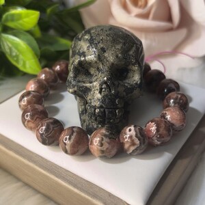 May include: A dark, metallic skull sculpture is centered, with a beaded bracelet of brown and cream-colored stones encircling it. The beads are round and patterned. The background includes a white surface and a soft pink rose.