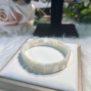 Peut inclure: Un bracelet blanc en nacre. Le bracelet est composé de perles carrées et iridescentes. Le bijou est exposé sur une surface blanche, avec d'autres bracelets visibles en arrière-plan. Le bracelet est de style extensible.