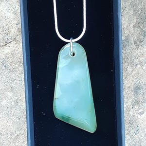 May include: A light green pendant necklace with a silver chain. The pendant is a smooth, rectangular shape with rounded edges and a hole at the top for the chain. The pendant is displayed in a black box.