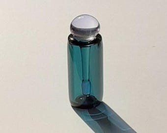 May include: A cylindrical teal glass bottle with a smooth, rounded, translucent white stopper. The bottle features a clear glass interior structure visible through the deep teal walls, creating a sleek, minimalist appearance with a polished finish.