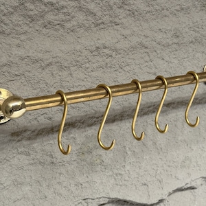 May include: A brass kitchen utensil rack with a round knob on the left end and seven hooks on the right. The rack is mounted to a wall.