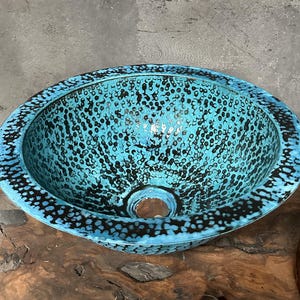 May include: A circular, turquoise sink with a speckled pattern of black dots. The sink has a wide rim with a dark border and a central drain hole. The sink is made of metal and is designed for bathroom use.