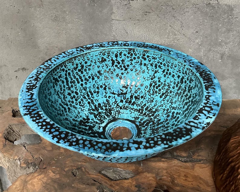 May include: A circular, turquoise sink with a speckled pattern of black dots. The sink has a wide rim with a dark border and a central drain hole. The sink is made of metal and is designed for bathroom use.