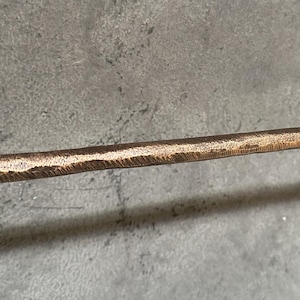 May include: A close-up of a hammered bronze metal rod. The rod has a textured surface and a slightly rounded edge.