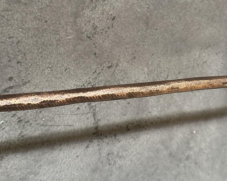 May include: A close-up of a hammered bronze metal rod. The rod has a textured surface and a slightly rounded edge.