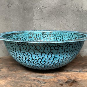 May include: A round, turquoise metal bowl with a textured surface and a dark speckled pattern. The bowl has a wide, flat rim and sits on a wooden surface.