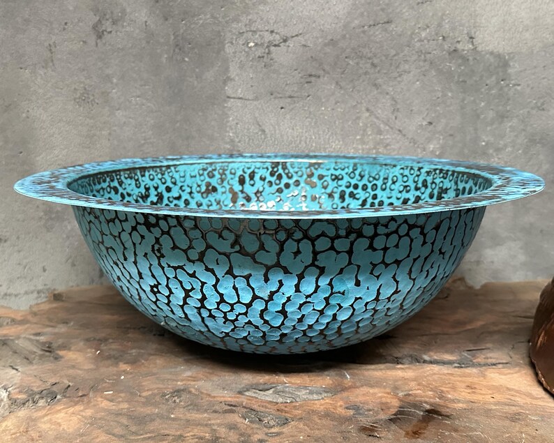May include: A round, turquoise metal bowl with a textured surface and a dark speckled pattern. The bowl has a wide, flat rim and sits on a wooden surface.