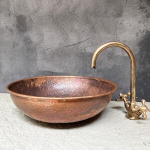 K&ouml;nnte beinhalten: Ein geh&auml;mmertes Kupferwaschbecken mit einem Messinghahn. Das Waschbecken ist oval geformt und hat eine strukturierte Oberfl&auml;che. Der Hahn hat einen einzigen Griff und einen Auslauf, der sich nach oben kr&uuml;mmt.
