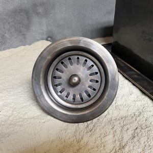 Könnte beinhalten: Ein runder, silberfarbener Spülbecken-Ablaufsieb mit einem kreisförmigen, perforierten Design. Das Sieb hat einen erhöhten Knopf in der Mitte.