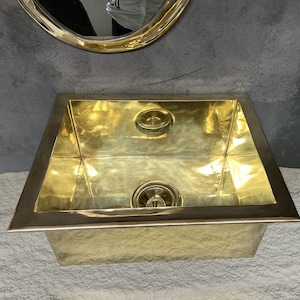 Puede incluir: Un lavabo de latón de color dorado, de forma rectangular redondeada, con un desagüe en el centro. El lavabo está colocado sobre un fondo gris.