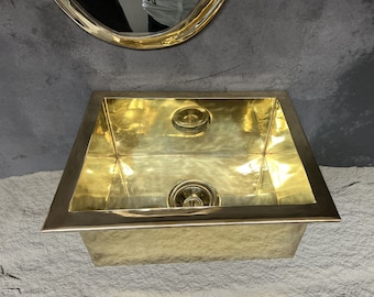 Puede incluir: Un lavabo de latón de color dorado, de forma rectangular redondeada, con un desagüe en el centro. El lavabo está colocado sobre un fondo gris.