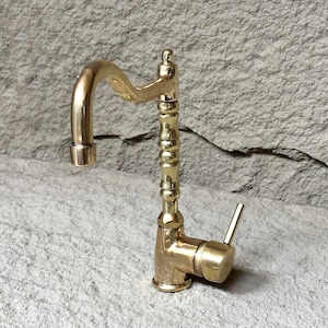 May include: A gold-toned kitchen tap with a curved spout and a decorative handle. The tap is mounted on a grey stone surface.