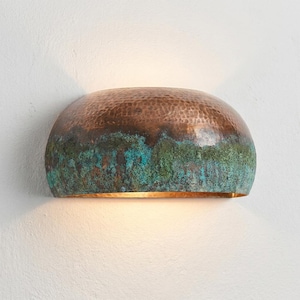 May include: A copper wall sconce with a hammered texture. The top is a rich brown, transitioning to green and blue hues at the bottom. The sconce emits a warm, inviting light, perfect for interior decor.