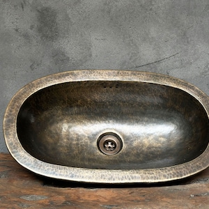 May include: An oval-shaped, hammered brass bathroom sink with a drain in the centre.