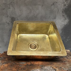 Puede incluir: Un fregadero de cocina cuadrado de latón martillado con un desagüe en el centro. El fregadero está sobre una superficie de madera.