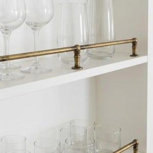 May include: A white cabinet with a brass bar and clear glassware. The cabinet has two shelves, with wine glasses and tumblers on display. The brass bar runs horizontally across the top shelf, adding a decorative touch to the barware.