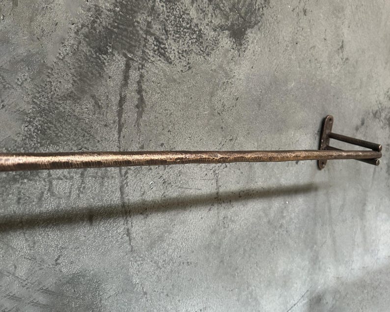 May include: A rustic, hammered copper towel bar with a simple design. The bar is mounted to a wall with two brackets.