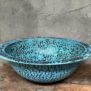 May include: A turquoise bowl with a speckled pattern and a dark rim. The bowl has a wide, shallow shape and sits on a wooden surface. The speckled pattern is black and covers the entire surface of the bowl.