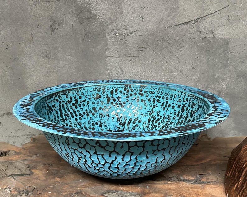 May include: A turquoise bowl with a speckled pattern and a dark rim. The bowl has a wide, shallow shape and sits on a wooden surface. The speckled pattern is black and covers the entire surface of the bowl.