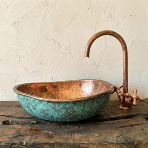 May include: A copper sink with a turquoise patina and a matching copper tap. The sink has an oval shape and sits on a wooden surface. The tap has a curved spout and cross-shaped handles.