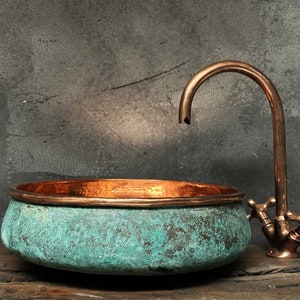 Può includere: Un lavandino in rame con una finitura in patina verde e un rubinetto in rame con due maniglie. Il lavandino è su una superficie in legno.