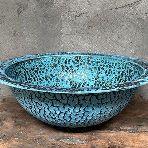 May include: A turquoise bowl with a textured surface and a black dotted pattern. The bowl has a wide rim and is set on a wooden surface. The bowl's interior is a lighter shade of turquoise.