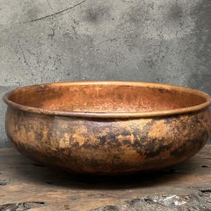 May include: A hammered copper bowl with a wide, shallow design. The bowl has a rich, aged patina with shades of brown and orange, and sits on a wooden surface. The interior and exterior show signs of wear, adding to its vintage appeal.