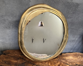 May include: A round, gold-toned, brass mirror with an irregular, wavy, organic shape. The mirror is reflecting a room with a light fixture.