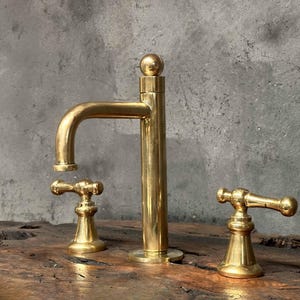 May include: A gold-toned bathroom faucet with two handles and a tall spout. The faucet is mounted on a wooden surface.