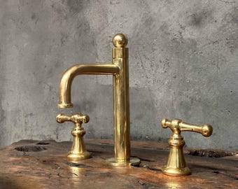 Könnte beinhalten: Ein goldfarbener Badezimmerhahn mit zwei Griffen und einem hohen Auslauf. Der Hahn ist auf einer Holzoberfläche montiert.