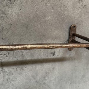May include: A rustic, brown metal towel bar with a hammered finish. The bar is mounted to a wall with a simple, rectangular bracket.