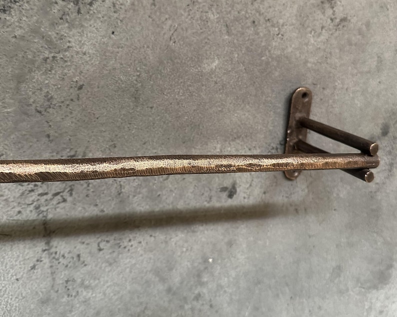 May include: A rustic, brown metal towel bar with a hammered finish. The bar is mounted to a wall with a simple, rectangular bracket.
