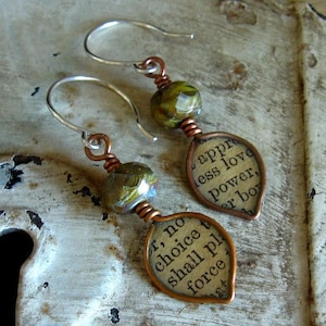 May include: A pair of earrings with copper wire and green glass beads. Each earring features a copper leaf charm with a printed quote. The left earring says "But no choice shall plead force" and the right earring says "...appre less love power...er boy".
