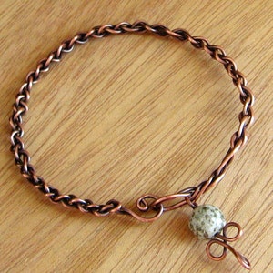 May include: A copper wire bracelet with a braided design and a single, speckled, light grey stone charm.