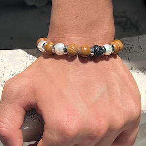 May include: A beaded bracelet featuring alternating round brown wood beads, smooth white pearls, and silver-toned metal spacers. A single textured black lava stone bead is positioned as a central focal point within the circular arrangement.