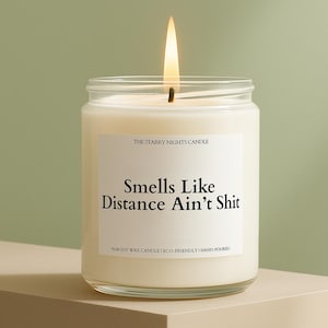 May include: A lit candle in a clear glass jar with a white label. The label reads "Smells Like Distance Ain't Shit" and "The Starry Nights Candle". The candle is on a beige surface against a green background.