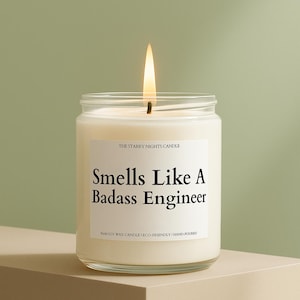 May include: A lit, white candle in a clear glass jar. The label reads "Smells Like A Badass Engineer." The candle is labeled "The Starry Nights Candle" and is made of soy wax. The flame is bright yellow.