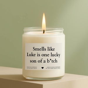 May include: A lit soy wax candle in a clear glass jar. The candle has a white label with the text "Smells like Luke is one lucky son of a b*tch". The candle is hand-poured in New Jersey.