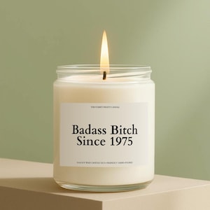 May include: A lit candle in a clear glass jar with a white label. The label reads "Badass Bitch Since 1975". The candle is on a neutral-toned surface against a green background. The text "THE STARRY NIGHTS CANDLE" is above the main text.