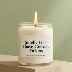 May include: A lit, white candle in a clear glass jar. The label reads "Smells Like I have Concert Tickets." The candle is on a neutral-toned surface against a green background. The text "100% Soy Wax Candle Eco-Friendly Hand-poured" is also visible.