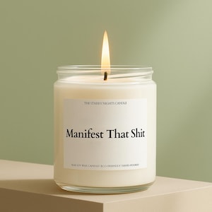 A white candle with a lit flame, labeled "Manifest That Shit" and "The Starry Night's Candle", sits on a beige surface against a green background.