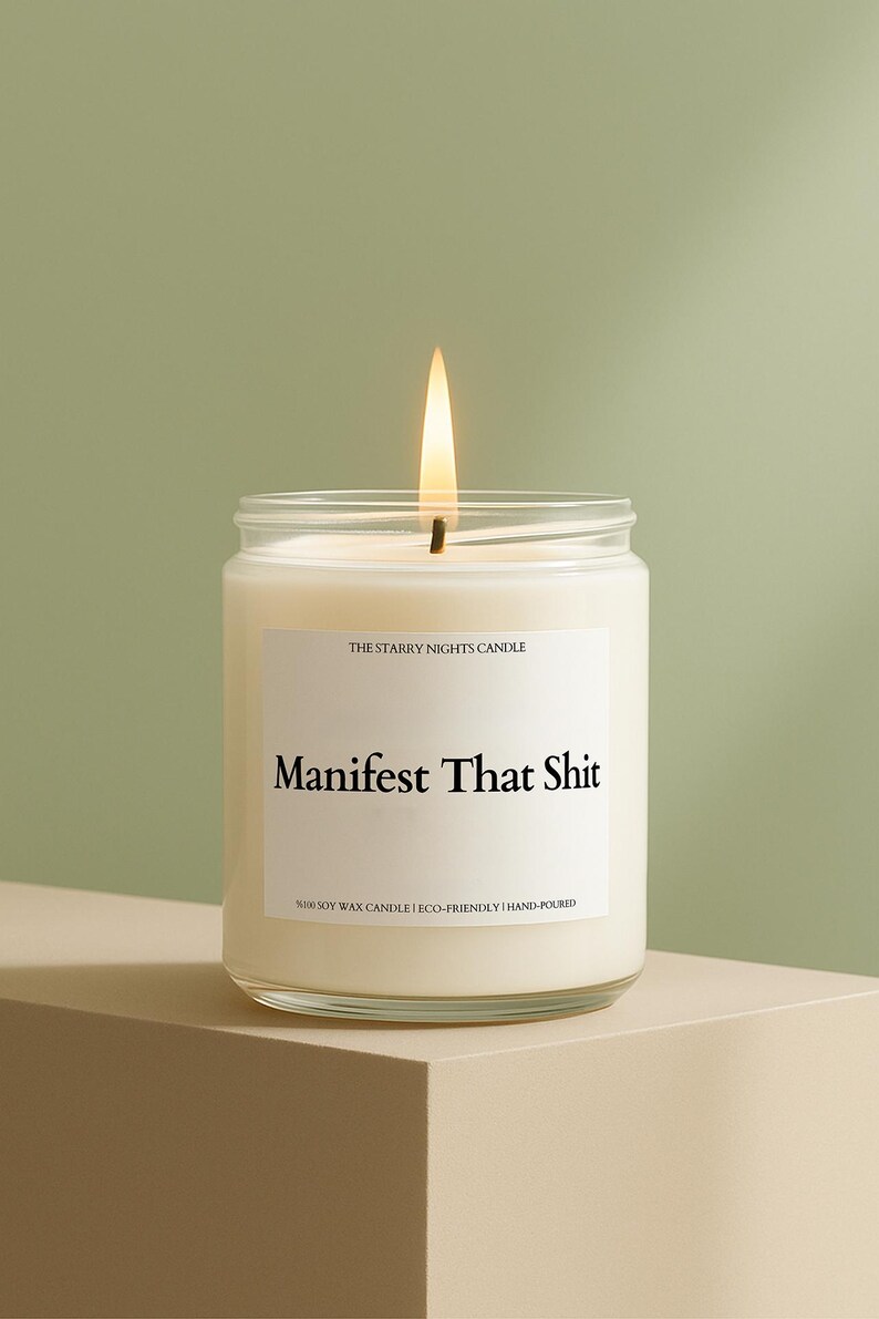 A white candle with a lit flame, labeled "Manifest That Shit" and "The Starry Night's Candle", sits on a beige surface against a green background.