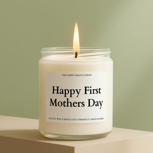 May include: A lit candle in a clear glass jar. The label reads "Happy First Mothers Day". The candle is made of soy wax and is hand-poured. The flame is bright against a soft green background.