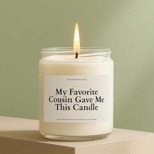 May include: A lit candle in a clear glass jar. The candle has a white label with the text "My Favorite Cousin Gave Me This Candle." The candle is on a neutral-toned surface against a green background.