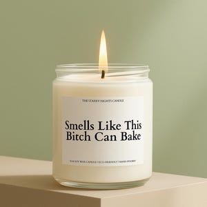 May include: A lit, white soy wax candle in a clear glass jar. The label reads "Smells Like This Bitch Can Bake." The candle is hand-poured and eco-friendly. The background is a soft green.