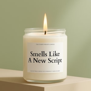 May include: A lit, white candle in a clear glass jar. The label reads "Smells Like A New Script" with "THE STARRY NIGHTS CANDLE" and "100% SOY WAX CANDLE | ECO-FRIENDLY | HAND-POURED" below. The flame glows brightly.